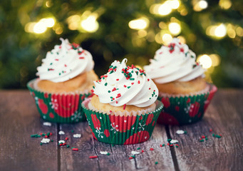 Christmas cupcakes with vanilla frosting and red and green sprinkles on rustic table. Shallow depth of field. Sparkling holiday lights background.