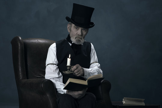 Dickens Scrooge Man Sitting in Chair with Book by Candlelight.