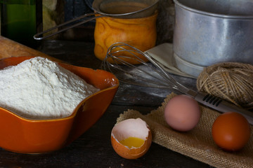 White wheat flour in ceramic ware, broken egg with the yolk, whole eggs and cooking utensils for cooking test. Rustic style. Side view close-up.