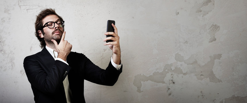 Portrait of man wearing trendy suit and makes selfe on his smartphone. Man stands against the concrete wall. Horizontal