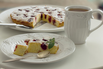 Tea in a white mug, a piece of cherry pie on a white plate on a white table in a range of natural daylight. Horizontal photo.