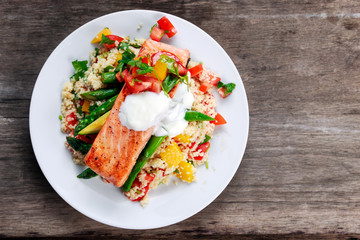 Pan fried Salmon with tender asparagus, courgette served on couscous mixed with sweet tomato, yellow pepper, with greek yogurt on old wooden table.