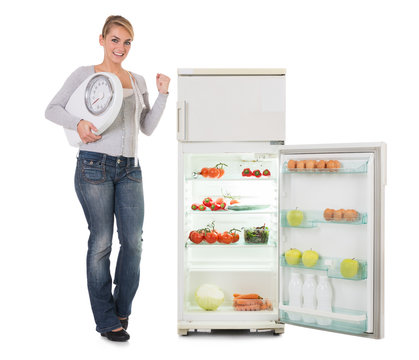 Woman Holding Weighing Scale While Standing By Refrigerator