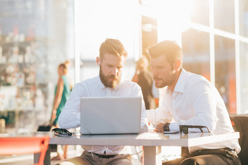 Two young bearded caucasian modern business man sitting in a bar, using smartphone and laptop, looking downward the screen, smiling - business, work, technology concept