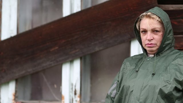 Senior woman in green raincoat stands near the boarded-up uninhabited house and looking at the camera
