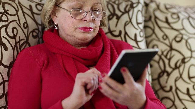 A senior woman sits on a sofa and uses electronic book. Woman with e-book

