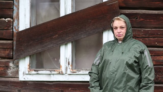 Woman in waterproof raincoat stands near the boarded-up uninhabited house and looks at the camera
