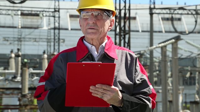 Equipment engineer on power plant. Worker in yellow hard hat at heat electric power station. Maintenance-mechanical engineer on outdoor switchgear. Engineering supervision. Man writes in a worksheet
