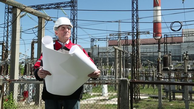 Maintenance staff with working drawings at nuclear power station. Worker in white helmet with engineering drawing near outdoor switchgear at atomic power station
