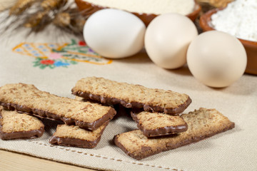 Rectangular cookies with chocolate in front of various ingredients such as wheat flour and eggs, on an embroidered cloth.