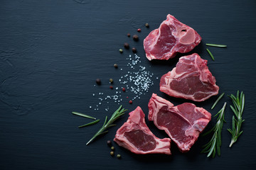Raw T-bone lamb steaks with seasonings on a black wooden surface