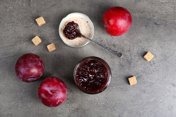 A jar of tasty jam, a spoon, plums and crackers on grey background