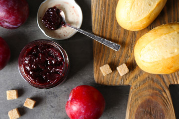 A jar of tasty jam, a spoon, plums, crackers on grey background and fresh buns on wooden tablet