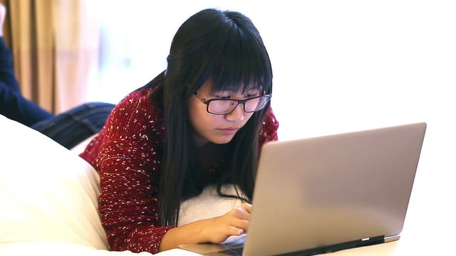 Asian Woman wearing glasses lying on a bed at home and she works on a laptop