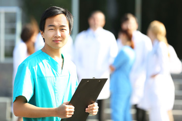 Young smiling doctor with clipboard in hands standing against group of medical workers