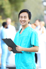Young smiling doctor with clipboard in hands standing against unfocused group of medical workers