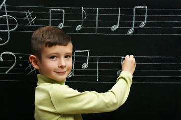 Cute boy writing at the blackboard with musical notes, in the classroom