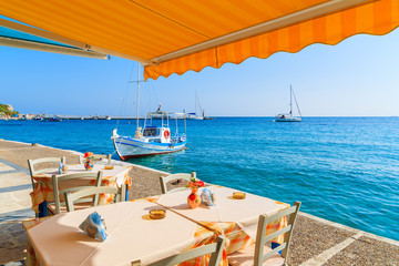 Table with chairs in Greek tavern on coast of Samos island with fishing boat in distance, Kokkari town, Greece
