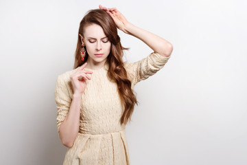 Attractive young woman in golden dress is standing, raising his hands to his face. Her gestures express doubts. She weares massive gold earrings.She has long brown hair.