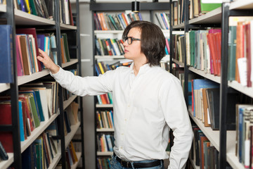 young man with dark hair choosing a book standing between shelve