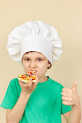 Little smiling boy in chefs hat is tasting the cooked pizza