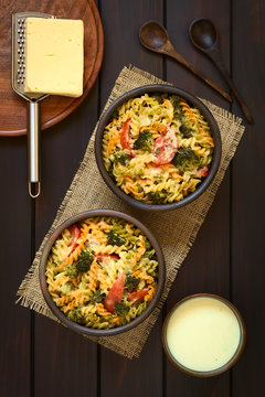 Baked Tricolor Fusilli Pasta And Vegetable (broccoli, Tomato) Casserole In Rustic Bowls, Cream Sauce, Spoons, Grater And Cheese On The Side, Photographed Overhead On Dark Wood With Natural Light