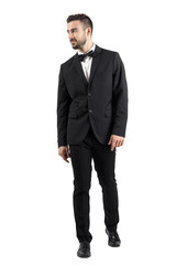 Young beaded man in tuxedo with bow tie walking towards the camera looking away. Full body length portrait isolated over white studio background.