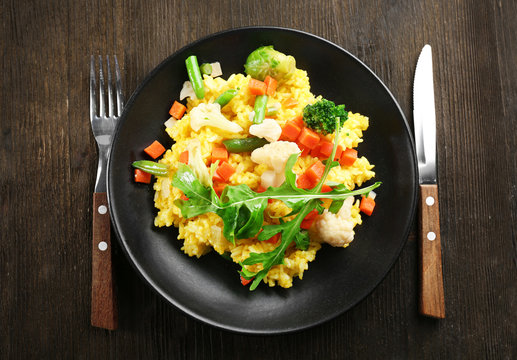 Black plate with vegetable risotto, fork and knife on wooden table