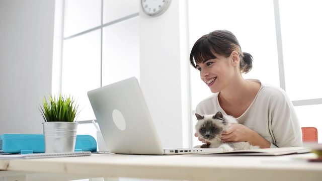 Smiling woman at home typing on a laptop and social networking, she is holding and cuddling her cat