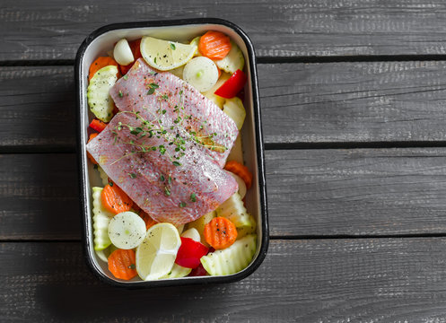 Cooking healthy food - raw ingredients: potatoes, zucchini, carrots, onions, garlic, peppers and fish sea bass in a baking dish on dark wooden surface