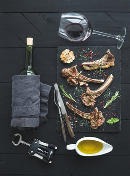 Grilled lamb chops. Rack of Lamb with garlic, rosemary, spices on slate tray, wine glass, oil in a saucer, cork screwer and bottle over black wood background