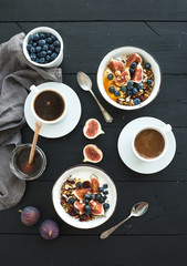 Obraz na płótnie Canvas Healthy breakfast set. Bowls of oat granola with yogurt, fresh blueberries and figs, coffee, honey, over black wooden backdrop