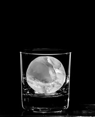A cocktail glass rimmed with light against a black background and a giant round ice ball, cube.
