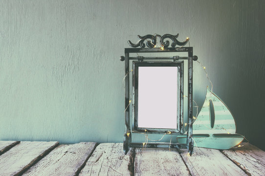 low key image of old victorian steel blue blank frame with fairy garland lights and wooden sailing boat on wooden table. vintage filtered image
