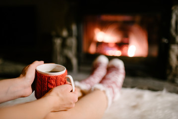 Woman holding a cup of tea by the Christmas fireplace. Woman relaxes by warm fire with cup of hot drink and warming up her feet in woollen socks. Close up on feet. Winter, Christmas holidays concept.