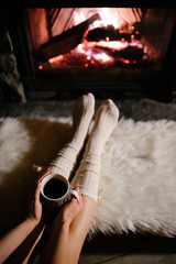 Feet in woollen socks by the Christmas fireplace. Woman relaxes by warm fire with a cup of hot drink and warming up her feet in woollen socks. Close up on feet. Winter and Christmas holidays concept