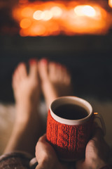 Bare woman feet by the cozy fireplace. Woman relaxes by warm fire with a cup of hot drink and warming up her feet. Close up on feet. Winter and Christmas holidays concept