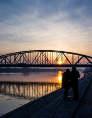 Couple walking on the boulevard of Vistula river in Poland with bridge on background. Torun, Poland - obrazy, fototapety, plakaty