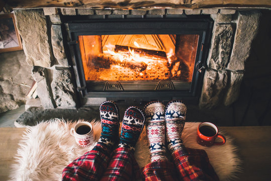 Feet in woollen socks by the Christmas fireplace. Family sitting relaxes by cozy authentic fireside with a cup of hot drink and warming up their feet. Winter and Christmas holidays concept