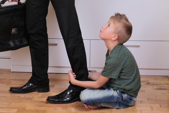 Boy holding dad's leg as he tries to leave for work. Child trying to stop father to go to work. Kid needs parent attention. Busy businessman father. Balance between work and time spent with family