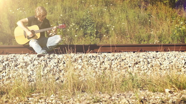 A long-haired guitarist sitting on the railway and playing