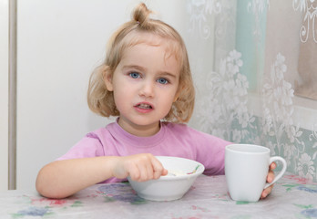 Little girl eats porridge from sitting at the table.