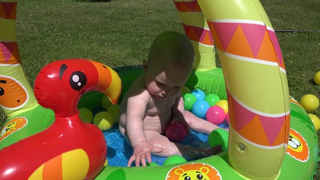 Cheerful laughing baby splash water first time in pool full of colorful balls on garden meadow. Natural true infant child emotions. Static shot. 4K 
