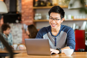 Happy cheerful asian male smiling and using laptop in cafe
