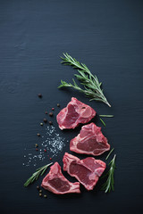 Raw T-bone lamb steaks with seasonings on a black wooden surface