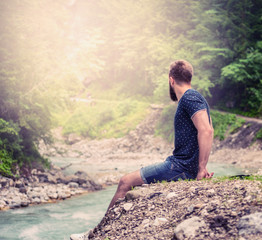 Naklejka premium young man with a beard and short hair, sitting on the bank of a mountain river in shorts and a blue shirt, with natural nature background