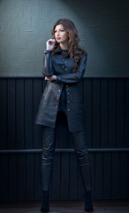 Charming young brunette woman in black leather outfit, coat and trousers, with dark gray wall on background. Sexy gorgeous young woman. Full length portrait of a sensual woman with long curly hair