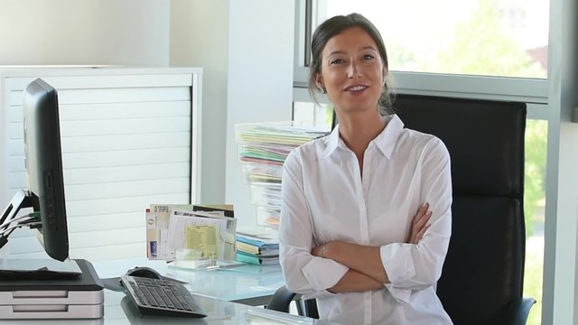 Businesswoman sitting at her desk and typing on her keyboard, she turns to the camera and say a few words, smiling and crossing his arms