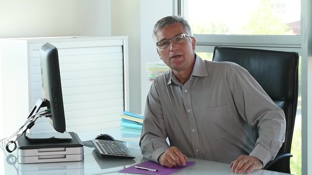 Senior businessman sitting at his desk and typing on his keyboard, he turns to the camera, smiling, and said a few words