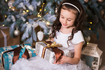 girl with curls and a smile sitting by a Christmas tree with a g